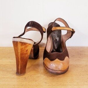 May include: A pair of brown suede heeled sandals with a snakeskin-patterned heel and toe cap. The sandals have an ankle strap with a buckle closure. The brand name "Balera" is printed on the insole.