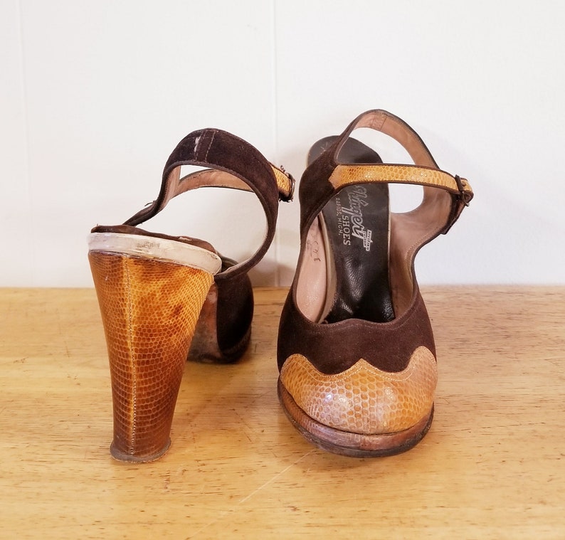 May include: A pair of brown suede heeled sandals with a snakeskin-patterned heel and toe cap. The sandals have an ankle strap with a buckle closure. The brand name "Balera" is printed on the insole.