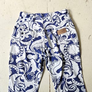May include: High-waisted, vintage Wrangler jeans with a bold blue and white floral pattern. The jeans feature a classic five-pocket design and a brown leather Wrangler patch on the back. The fabric is a sturdy denim.