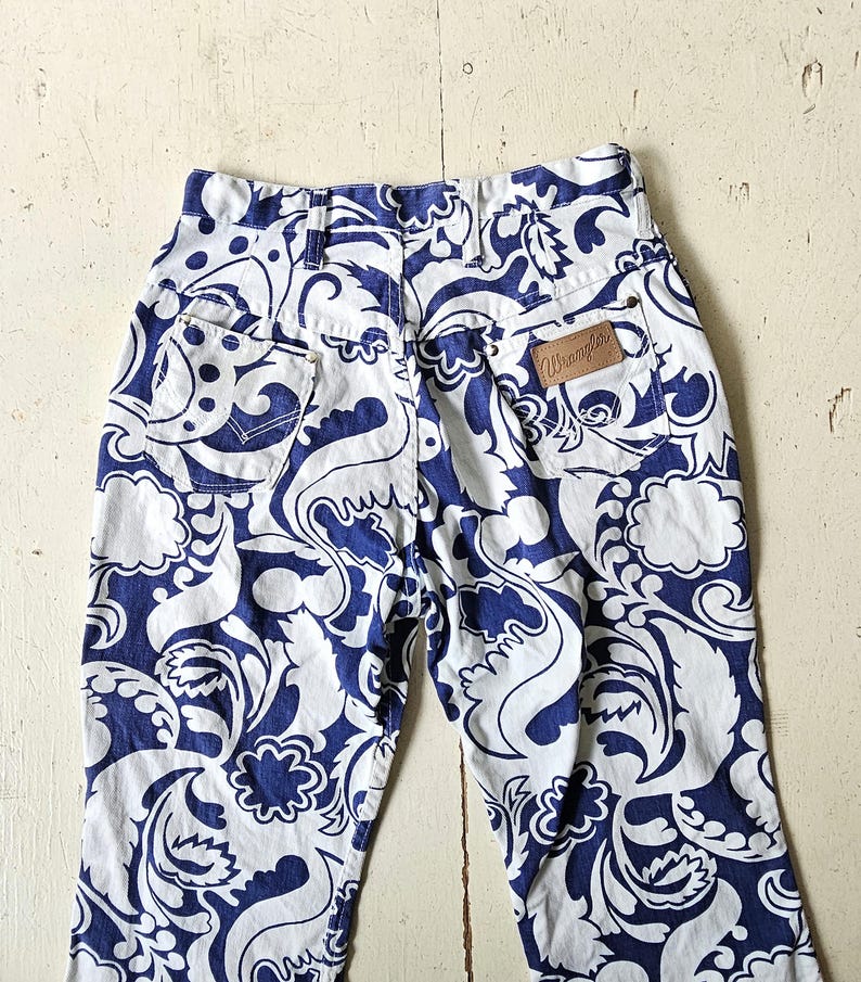 May include: High-waisted, vintage Wrangler jeans with a bold blue and white floral pattern. The jeans feature a classic five-pocket design and a brown leather Wrangler patch on the back. The fabric is a sturdy denim.