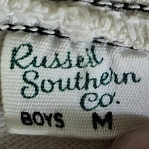 May include: Close-up of a white fabric tag with the text "Russell Southern Co." in green script. Below, the tag reads "BOYS M" in black block letters. The tag is sewn onto a white, textured fabric.