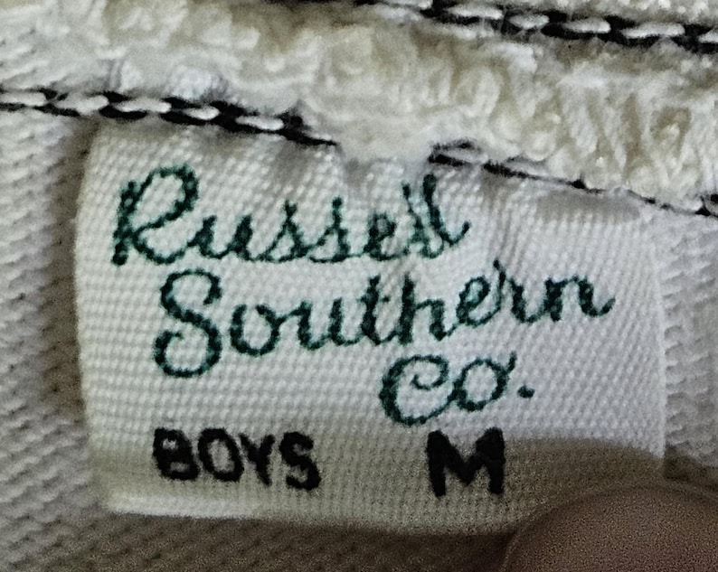 May include: Close-up of a white fabric tag with the text "Russell Southern Co." in green script. Below, the tag reads "BOYS M" in black block letters. The tag is sewn onto a white, textured fabric.