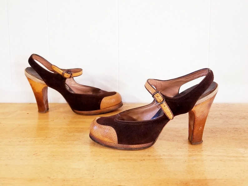 May include: A pair of brown suede heeled shoes with a tan leather toe cap and a buckle strap. The shoes have a chunky wooden heel.