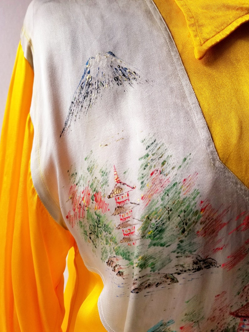 Puede incluir: Una camisa blanca con cuello amarillo y mangas largas. La camisa tiene un dise&ntilde;o de inspiraci&oacute;n japonesa con una monta&ntilde;a, &aacute;rboles y una pagoda impresos en la parte delantera.