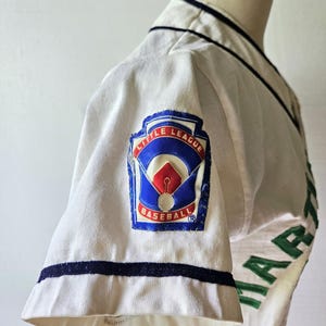 May include: A vintage baseball jersey, white with navy blue trim and green lettering. The jersey features a "Little League Baseball" patch on the sleeve, with red, white, and blue colors. The jersey is displayed on a mannequin.