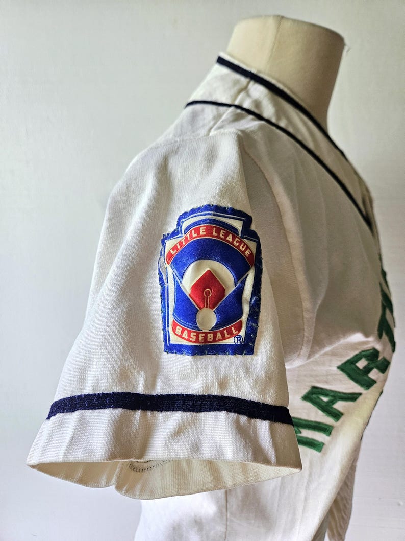 May include: A vintage baseball jersey, white with navy blue trim and green lettering. The jersey features a "Little League Baseball" patch on the sleeve, with red, white, and blue colors. The jersey is displayed on a mannequin.