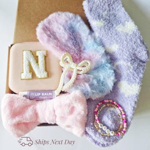 May include: A gift box containing a pink case with a gold letter "N", a lip balm, a pink bow, a white bow-shaped hair clip, a pair of fuzzy mittens, a pair of lavender socks with white cloud patterns, and a beaded bracelet. The box is filled with self-care items.
