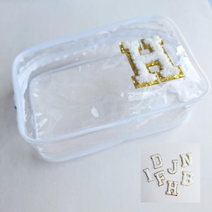 Puede incluir: Un neceser rectangular transparente con cremallera blanca y un parche con la letra "H" dorada y blanca. Se ven parches de letras doradas y blancas adicionales en la esquina inferior derecha. La bolsa está vacía.