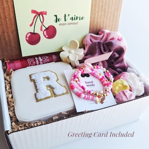 May include: A gift box filled with various items, including a white box with a gold glitter letter "R", a pink and white beaded bracelet, a pink scrunchie, a pink lip gloss, and a white flower-shaped hair clip. The box also includes a greeting card with the text "Je t'aime mon amour" in French.