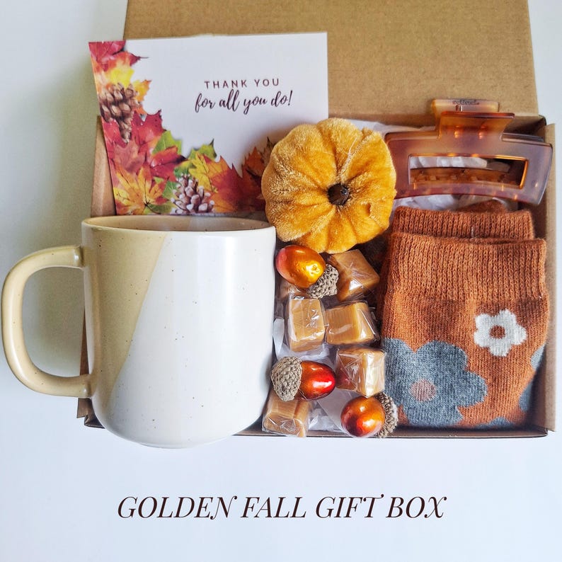 May include: A fall-themed gift box with a beige and white mug, caramel candies, a pumpkin decoration, a hair clip, and orange socks with a floral design. A card with fall leaves and the text "THANK YOU for all you do!" is also included. The box is labeled "GOLDEN FALL GIFT BOX".