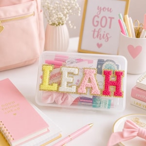May include: A clear rectangular plastic storage case featuring the name LEAH in large, textured, multi-colored varsity-style letters with gold glitter borders. The case is filled with various stationery items, including pink pens, hair accessories, and small containers.