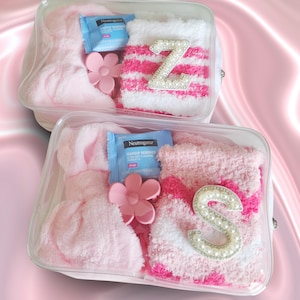 Peut inclure: Deux trousses transparentes zippées contenant des chaussettes duveteuses roses et blanches avec des lettres Z et S ornées de perles, une pince à cheveux rose en forme de fleur et un sachet de lingette démaquillante Neutrogena.