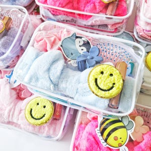 May include: A clear rectangular plastic storage case containing a light blue plush headband, a yellow fuzzy smiley face patch, a blue flower-shaped hair claw clip, a small bee-themed sticker, and a cosmetic tube with a metallic gold cap.