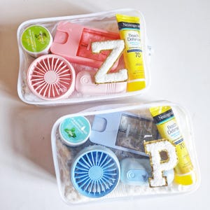 May include: Two clear travel bags filled with various items. One bag contains a pink fan, a pink razor, a tube of sunscreen, a lip sleeping mask, and a letter "Z". The other bag has a blue fan, a tube of sunscreen, a lip sleeping mask, and a letter "P".