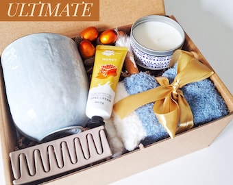 May include: A cardboard gift box containing a light blue ceramic mug, a tube of honey hand cream, a candle in a patterned tin, a pair of fuzzy blue and white socks tied with a gold ribbon, and a matte mauve hair claw clip with the text ULTIMATE.