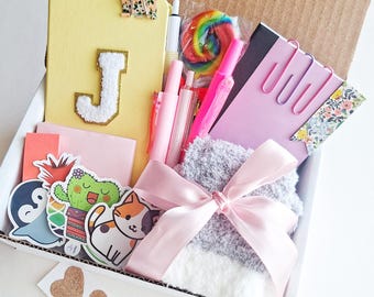 May include: A cardboard gift box containing a yellow notebook with a white letter J, a pink notepad with three pink paperclips, a rainbow lollipop, several pink pens, a pair of gray fuzzy socks tied with a pink ribbon, and various stickers.