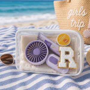 May include: A clear rectangular zipped pouch containing a light purple handheld fan, a lavender-coloured tube, a small round yellow container labelled Lip Sleeping Mask, and a white letter R patch with a gold glitter border, all resting on a white cloth.