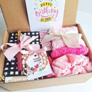 Puede incluir: Una caja de regalo llena de artículos rosas y blancos, que incluyen una pulsera, bálsamo labial, calcetines y un cuaderno. También se incluye una tarjeta con las palabras "Happy Birthday to You". La caja está atada con una cinta rosa.