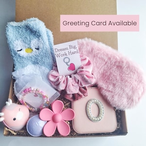 Pode incluir: Uma caixa de presente cheia de vários itens, incluindo uma máscara para os olhos de pelúcia rosa, um scrunchie de veludo rosa, uma meia de pantufa de pelúcia azul, uma bomba de banho rosa, um brinquedo de banho rosa em forma de flor, um chaveiro rosa com um charme de coração, uma pulseira de contas rosa e branca e um espelho compacto rosa com uma letra "O" incrustada de pérolas. A caixa também inclui um cartão com o texto "Dream Big, Work Hard".
