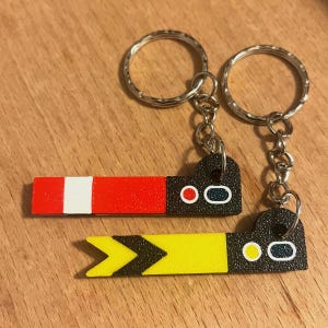 May include: Two keyrings featuring a black, heart-shaped design and a rectangular bar. One bar is red with a white stripe, the other is yellow with black chevrons. Each keyring has a silver ring.