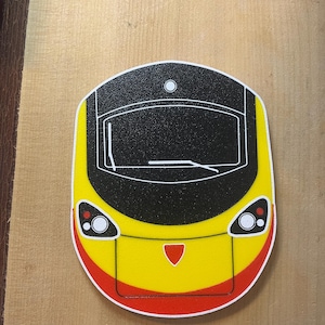 May include: A coaster featuring a stylized train front design. The coaster has a yellow base with a black top section, red accents, and white details. The design includes headlights, a windshield, and a red shield.