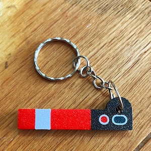 May include: A red and black keychain with a white stripe and a silver key ring. The keychain has a rectangular design with a black end featuring two red circles and a white oval.