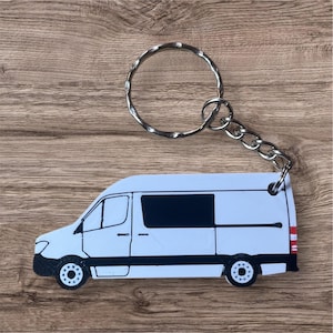 May include: A white van-shaped keychain with black detailing and a silver keyring. The van has a black window and black wheels. The keychain is on a wooden surface.