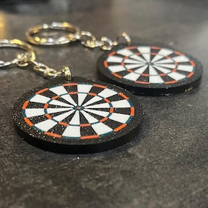 May include: Two dartboard keychains. Each keychain features a circular dartboard design with a black border, white and black sections, and orange and teal accents. The keychains are attached to silver keyrings.