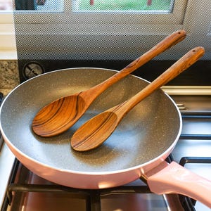 May include: A pink frying pan with a gray interior and two wooden spoons. The spoons are made of a light-colored wood with a visible grain. The pan is on a gas stove, and a small potted plant is visible in the background.