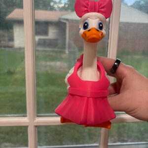 May include: A whimsical white goose figurine with a pink bow, dress, and accents. The goose has an orange beak and feet, and blue eyes. The figurine is held in front of a window.