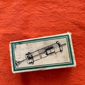 May include: A rectangular off-white cardboard box featuring a dark green border and a black line drawing of a medical syringe centered on the front. The syringe illustration includes clear measurement markings and the text ccm printed on the barrel.