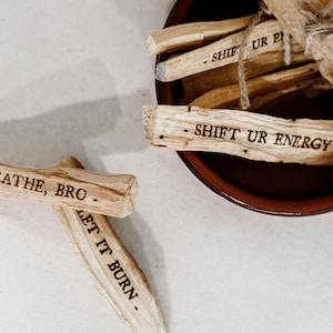 May include: Four wooden sticks with inspirational messages burned into them. The messages are "Breathe, Bro", "Let It Burn", "Shift Ur Energy", and "Shift Ur Energy". The sticks are in a brown bowl.