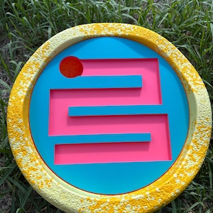 May include: A circular, yellow-painted wooden disc with a blue and pink maze design. The maze has a red circle at the top and a series of horizontal and vertical lines.