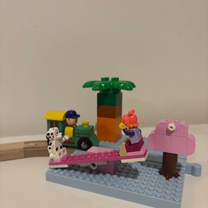 Könnte beinhalten: Ein Lego-Spielset mit einem Dalmatiner, einem Zug und zwei Lego-Figuren auf einer rosa Wippe. Ein rosa Baum und ein grüner und orangefarbener Turm sind ebenfalls vorhanden, alles auf einer blauen Basis.