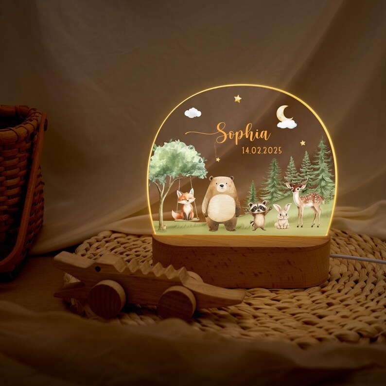 May include: A personalized night light featuring a woodland scene with a bear, fox, raccoon, rabbit, and deer. The name "Sophia" and the date "14.02.2025" are displayed on the light. A wooden alligator toy is in the foreground.