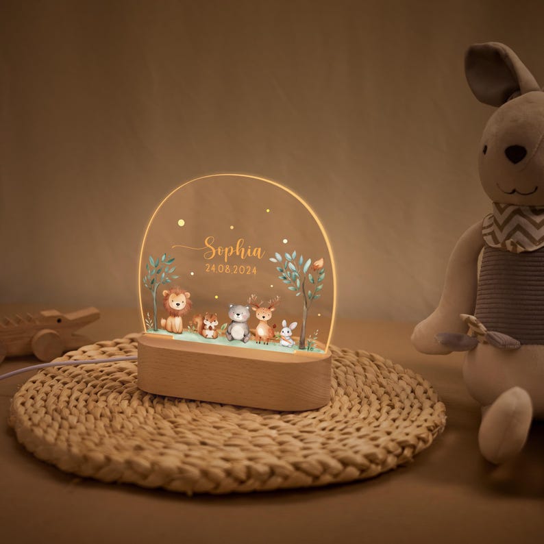 Pode incluir: Luz noturna personalizada com base em madeira e painel acr&iacute;lico transparente arqueado. O painel exibe uma cena de floresta com ilustra&ccedil;&otilde;es de animais, o nome "Sophia" e a data "24.08.2024". Ideal para o quarto de uma crian&ccedil;a.