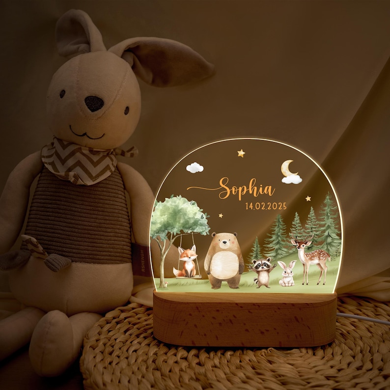 May include: A personalized night light featuring a woodland scene with a fox on a swing, a bear, raccoon, rabbit, and deer. The name "Sophia" and the date "14.02.2025" are displayed on the acrylic panel, set on a wooden base.