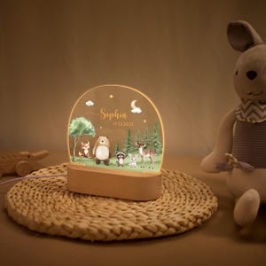 May include: A decorative night light featuring a woodland scene with a fox, bear, raccoon, rabbit, and deer. The light has a wooden base and a clear acrylic panel with the name "Sophia" and the date 14.02.2025. The light is on a woven mat.