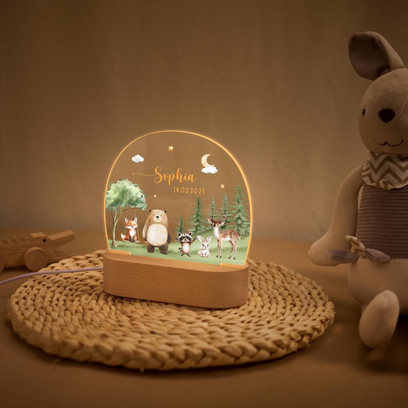 May include: A decorative night light featuring a woodland scene with a fox, bear, raccoon, rabbit, and deer. The light has a wooden base and a clear acrylic panel with the name "Sophia" and the date 14.02.2025. The light is on a woven mat.