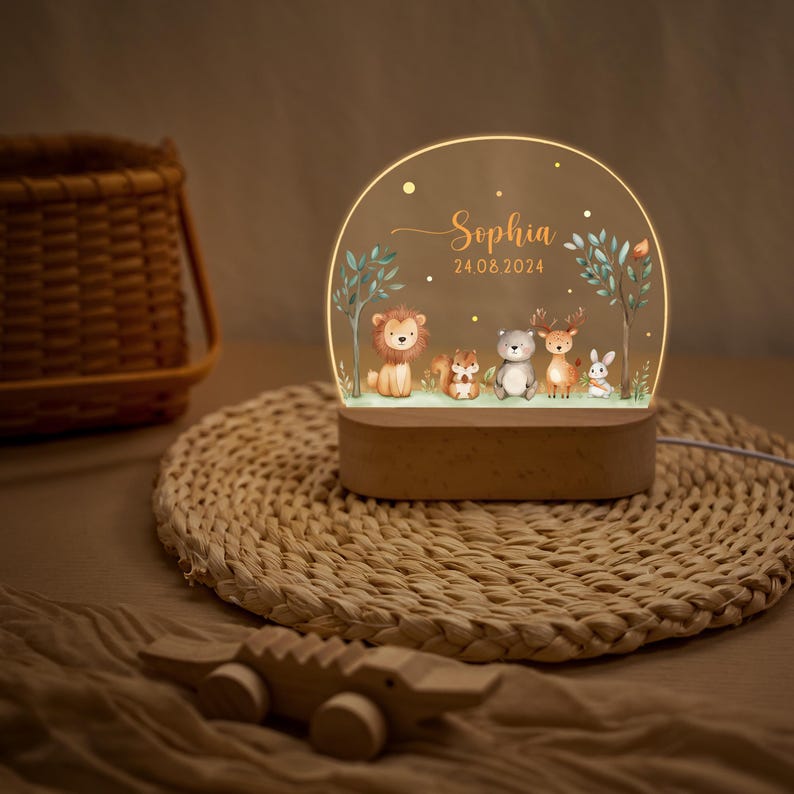 Pode incluir: Uma luz noturna decorativa com uma cena da floresta que apresenta um le&atilde;o, um esquilo, um urso, um veado e um coelho. O painel acr&iacute;lico transparente &eacute; iluminado e apresenta o nome "Sophia" e a data 24.08.2024. A base &eacute; de madeira.