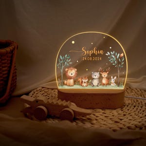 Pode incluir: Uma luz noturna personalizada com uma base de madeira e um painel acr&iacute;lico semicircular. O painel exibe uma cena de floresta com um le&atilde;o, um esquilo, um urso, um veado e um coelho. O nome "Sophia" e a data 24.08.2024 tamb&eacute;m s&atilde;o exibidos.
