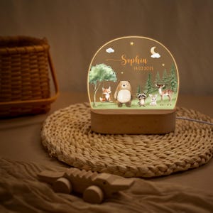 May include: A decorative night light featuring a woodland scene with a fox on a swing, a bear, raccoon, rabbit, and deer. The light has a wooden base and a clear acrylic panel with the name "Sophia" and the date 14.02.2025.