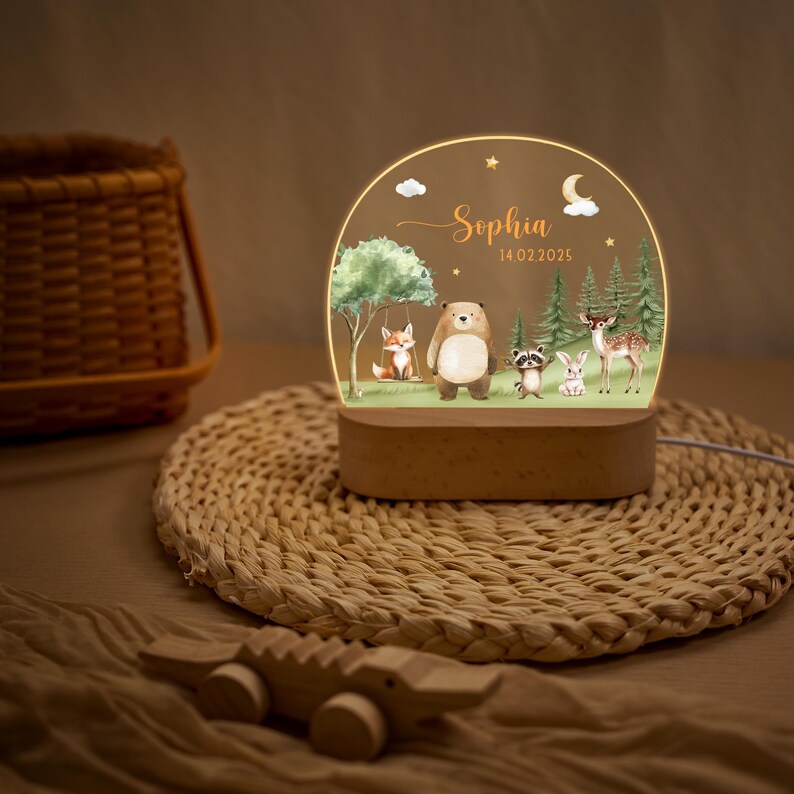 May include: A decorative night light featuring a woodland scene with a fox on a swing, a bear, raccoon, rabbit, and deer. The light has a wooden base and a clear acrylic panel with the name "Sophia" and the date 14.02.2025.