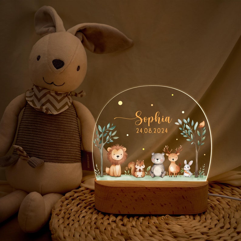 Pode incluir: Uma luz noturna personalizada com uma base de madeira e um painel de acr&iacute;lico transparente. O painel apresenta uma cena de floresta com um le&atilde;o, um esquilo, um urso, um veado e um coelho. O nome "Sophia" e a data "24.08.2024" tamb&eacute;m s&atilde;o exibidos.