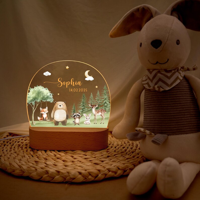 May include: A personalized night light with a wooden base and a clear acrylic panel. The panel features a forest scene with a fox, bear, raccoon, rabbit, and deer. The name "Sophia" and the date "14 02 2025" are also displayed. A plush bunny is in the background.