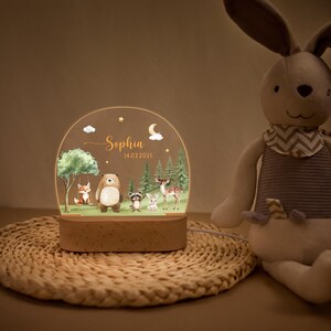 May include: A personalized night light featuring a woodland scene with a fox, bear, raccoon, rabbit, and deer. The acrylic panel is illuminated by a wooden base, with the name "Sophia" and date 14.02.2025.