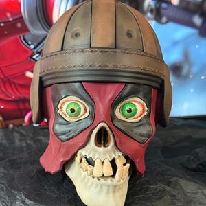 Könnte beinhalten: Eine rote und schwarze Totenkopfmaske mit grünen Augen und einem braunen Helm. Die Maske ist aus einem harten Material gefertigt und hat ein detailliertes Design.