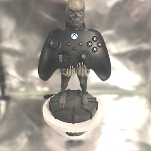 Könnte beinhalten: Ein schwarzer Videospiel-Controller, gehalten von einer grauen, skelettartigen Figur mit einem schädelartigen Gesicht. Die Figur steht auf einem schwarzen runden Sockel.