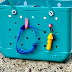 May include: A teal blue Bogg Bag with white polka dots and a white handle. The bag has a pink hook on each side and a yellow whistle attached to a carabiner on the right side. A blue swimming goggle is hanging on the left hook.