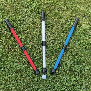 May include: Three golf alignment sticks in red, white, and blue, with black handles and bases, are arranged on a green grassy surface. A white golf ball rests near the sticks. The sticks are designed for golf training.